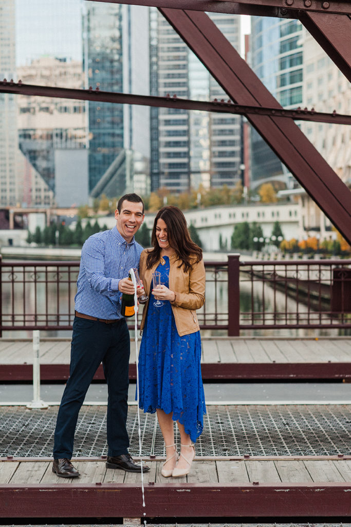 Couple pops open bottle of champagne on Kinzie Street Bridge for Chicago engagement photo