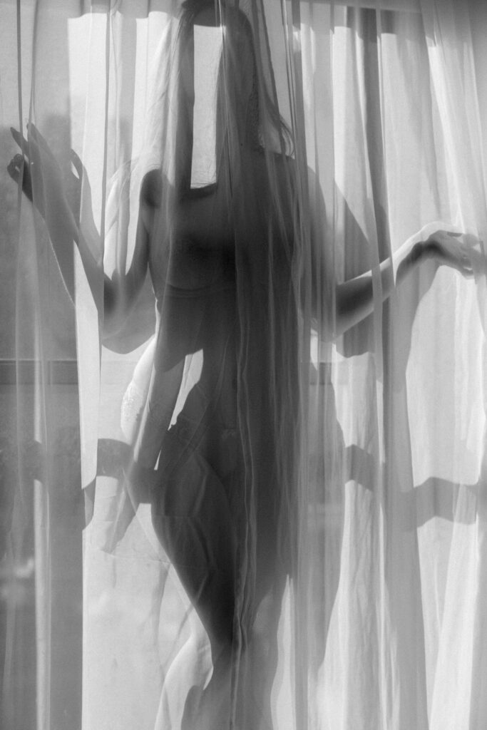 Boudoir image of a woman behind sheer curtains creating a soft silhouette