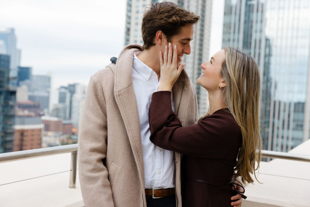 Newly engaged couple smiling at each other on rooftop terrace with city skyline behind them.