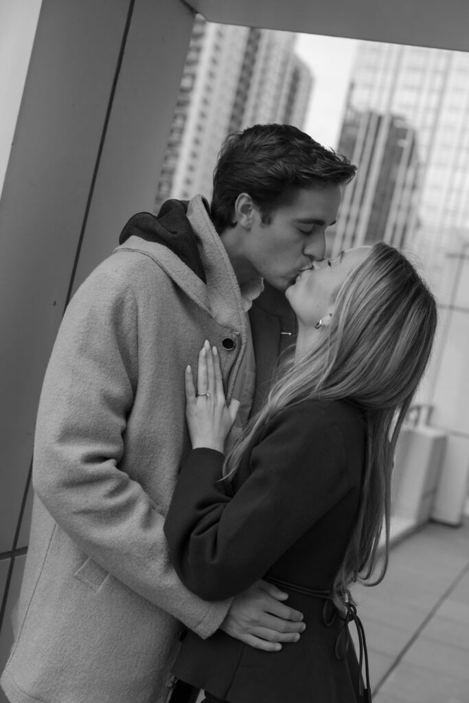 Black and white portrait of couple kissing on rooftop terrace after proposal.