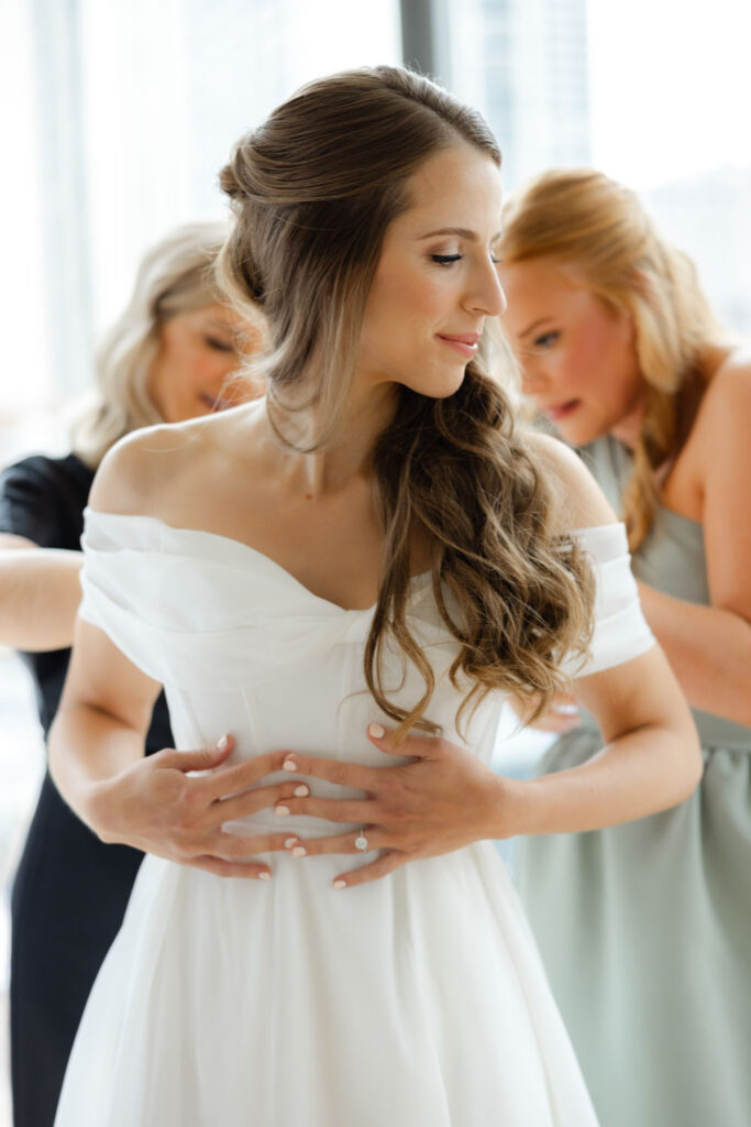 Bride being helped into her off-the-shoulder wedding gown by a bridesmaid