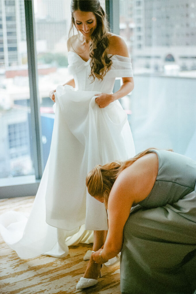 Bridesmaid helping adjust bride’s shoe while she stands near a hotel window caught on film