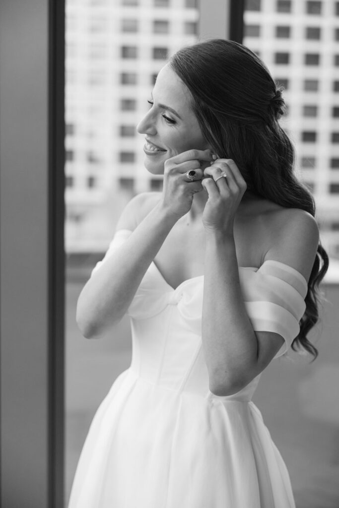 Bride putting on earrings beside a window in black and white portrait