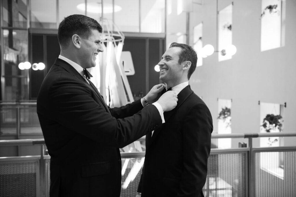 Groom adjusting boutonniere with help from a friend indoors