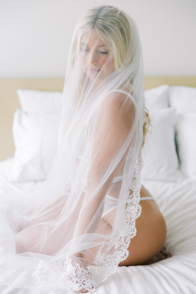 Bride wearing wedding veil sitting on bed in white lingerie soft natural light