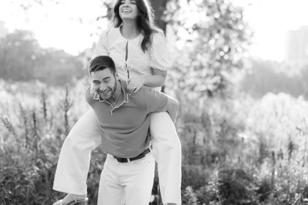 Couple laughing with piggyback ride in grassy Lincoln Park field black and white engagement photo