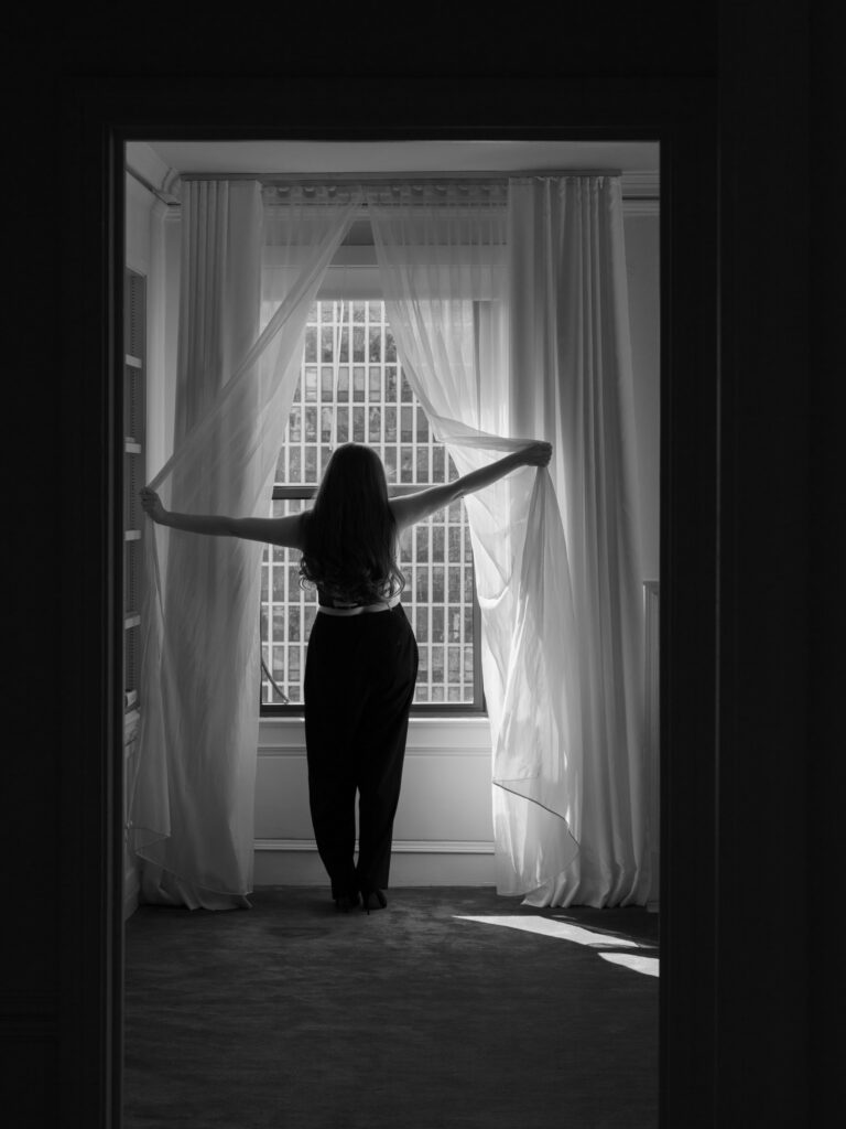 Black and white boudoir silhouette of woman opening sheer curtains at window