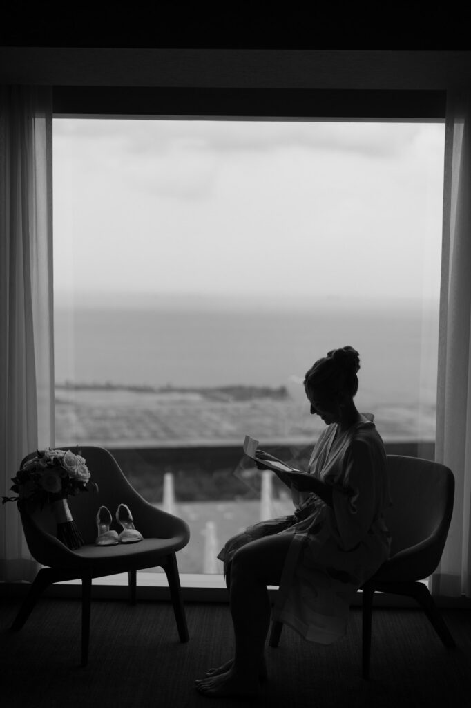 Bride sitting by the window in soft light reading a note before getting dressed on her wedding day.