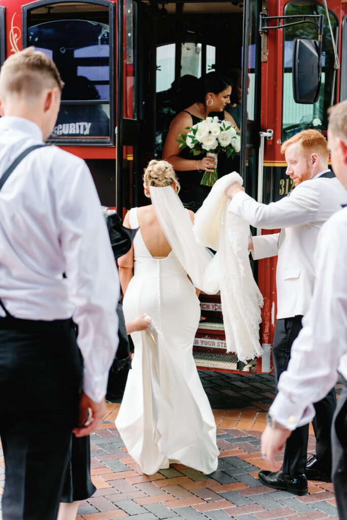 Bride stepping onto a trolley with her dress as wedding party members help her aboard.