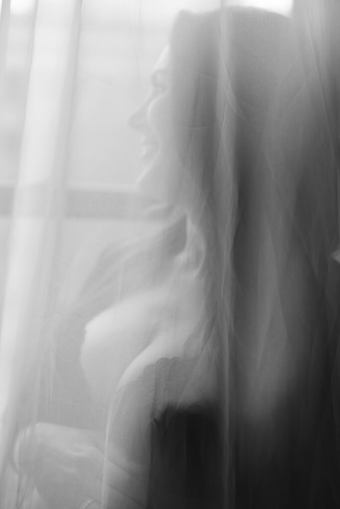 Black and white maternity boudoir image of woman behind sheer curtains