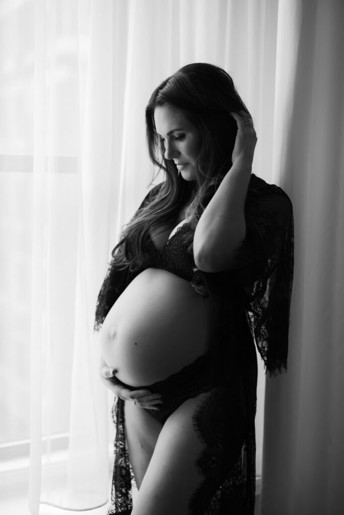 Black and white maternity portrait of pregnant woman standing by sheer curtains