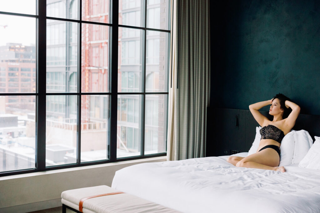 Wide view of hotel room with floor to ceiling windows and woman on bed in lingerie