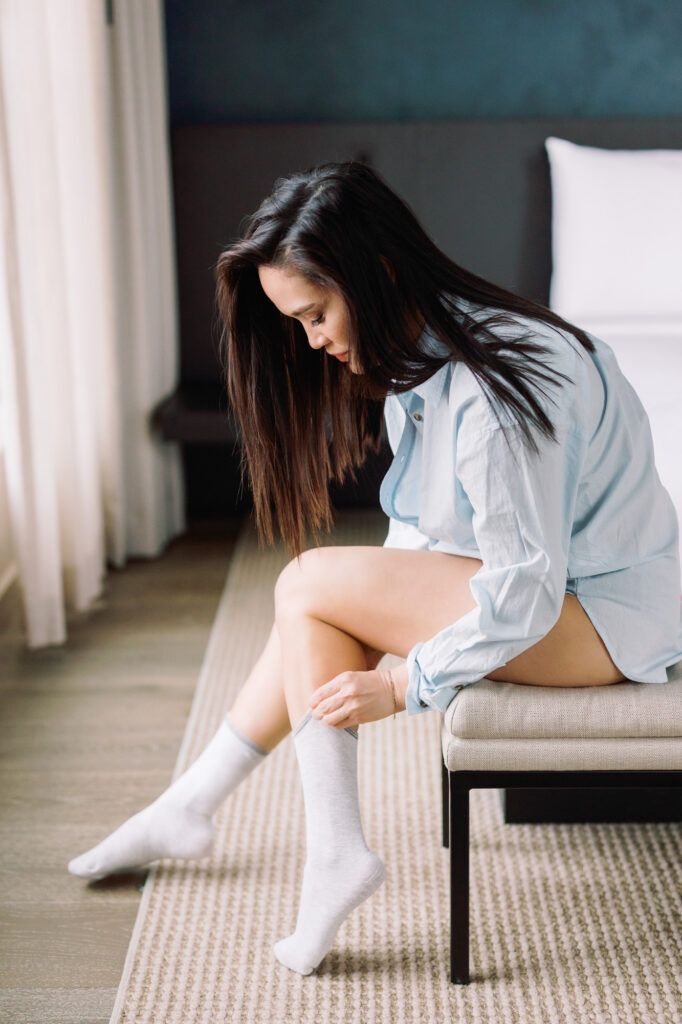Woman in oversized light blue button down sitting on bench adjusting cozy socks by window