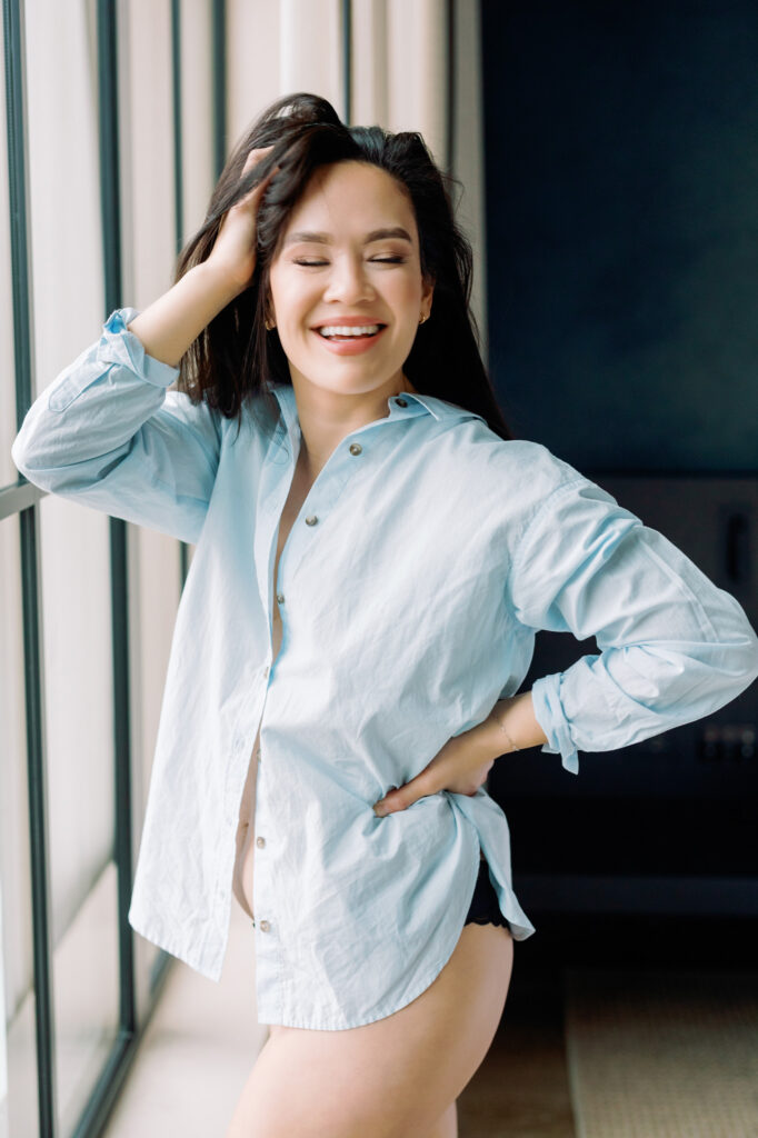 Woman smiling in button down shirt near window, relaxed boudoir moment